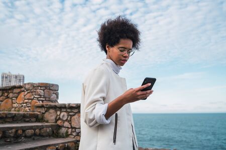 Portrait of young beautiful latin woman using her mobile phone while walking on coast line. Technology and communication concept.の写真素材