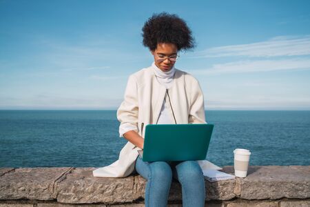 Portrait of young latin woman using her laptop while sitting against the sea. Technology concept.の写真素材