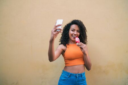 Portrait of afro woman taking selfies with her mophile phone while eating ice-cream. Technology and...