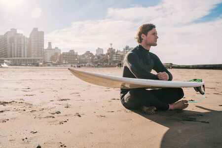 Young surfer sitting on sandy beach looking at the ocean with his surfboard. Sport and water sport concept.の写真素材