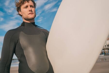 Close-up of young surfer standing at the beach with his surfboard and wearing black surfing suit. Sport and water sport concept.の写真素材