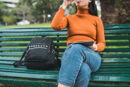 Portrait of young Asian woman using her digital tablet while drinking a cup of coffee in the park outdoors.の写真素材