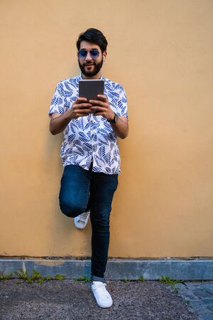 Portrait of young latin man using his digital tablet with earphones against yellow wall. Technology and urban concept.の写真素材