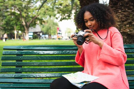 Portrait of young beautiful afro american woman using a professional camera while holding a map in a park. Travel concept.の写真素材