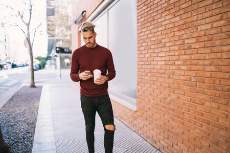 Portrait of young handsome man using his mobile phone while holding a cup of coffee outdoors in the street. Communication concept.の写真素材