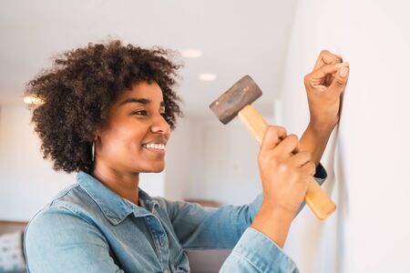 Portrait of young afro woman hammering nail on the wall at home. Stay home for renovating house. Lock down and Self-quarantine at home. Covid-19.の写真素材