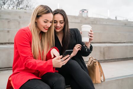 Portrait of two young friends using their mobile phone while sitting outdoors at the street....