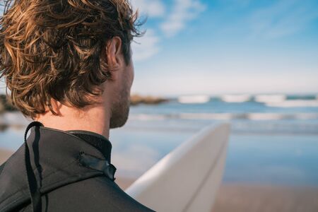 Young surfer standing at sandy beach with his surfboard while looking at the sea. Sport and water sport concept.の写真素材