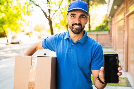 Portrait of a delivery man holding package and showing mobile phone. Delivery service and shipping concept.の写真素材