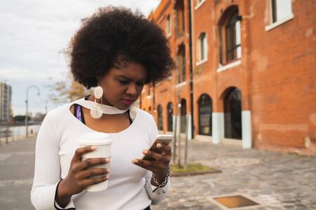 Portrait of Afro latin woman using her mobile phone while holding a cup of coffee outdoors in the...