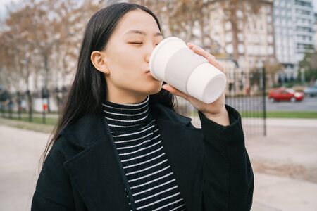 Portrait of young beautiful Asian woman drinking a cup of coffee and standing outdoors in the street. Urban concept.の写真素材