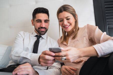 Portrait of two business people taking a break from work, lying on bed and using mobile phone at the...