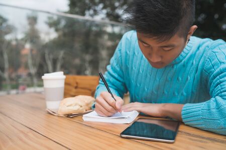 Portrait of young Asian man studying with his digital tablet and taking some notes while sitting in a coffee shop. Technology concept.の写真素材