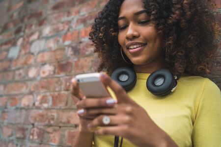 Portrait of young Afro american woman with headphones and using her mobile phone in the street....