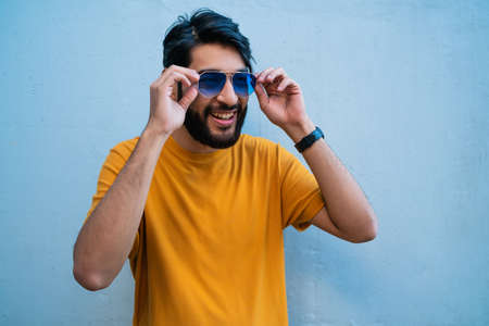 Portrait of young handsome man wearing summer clothes and sunglasses against blue background.