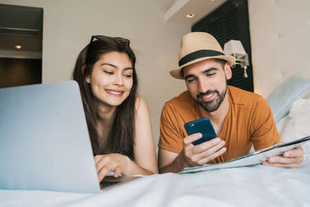 Portrait of lovely couple organizing their trip with laptop and mobile phone at the hotel room. Travel and holiday concept.の写真素材
