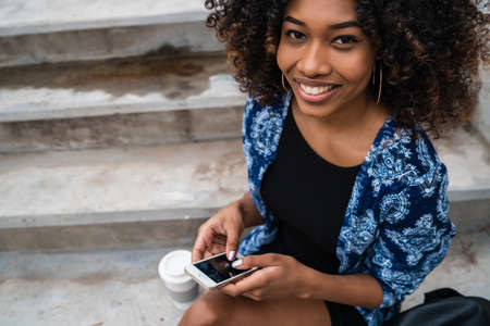 Portrait of young beautiful afro american woman using her mobile phone while sitting on concrete steps outdoors.の写真素材