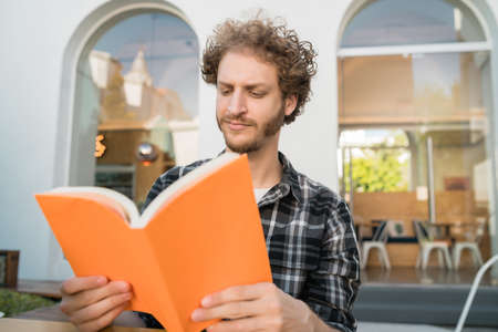 Portrait of caucasian man enjoying free time and reading a book while sitting outdoors at coffee shop.の写真素材