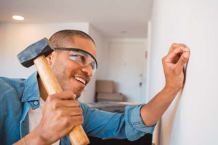 Portrait of young latin man hammering nail on the wall at home. Home improvement and repair home...