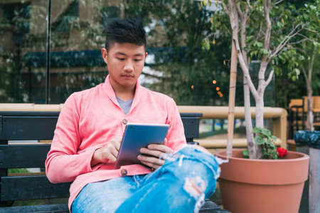 Portrait of young Asian man using his digital tablet while sitting in a bench outdoors. Technology...