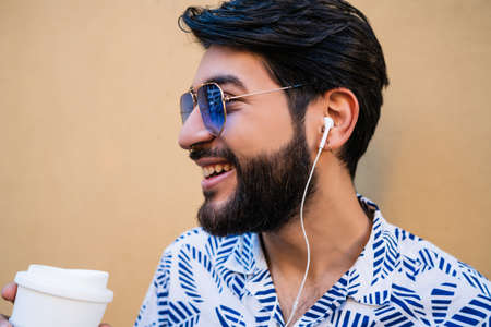 Portrait of young latin man wearing summer clothes, holding a cup of coffee and listening to music with earphones against yellow background.の写真素材