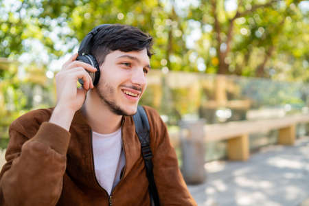 Portrait of young handsome man listening to music with headphones while sitting outdoors. Urban...