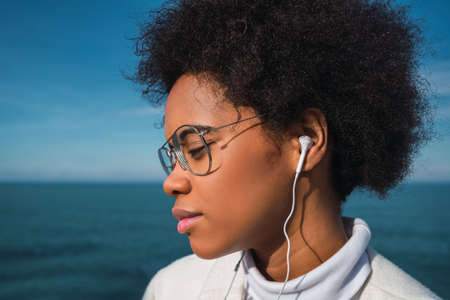Portrait of young latin woman listening to music with earphones with the sea on background. Music,...
