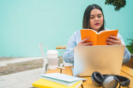 Portrait of young pluse size woman studying with laptop and books while sitting outdoors at coffee...