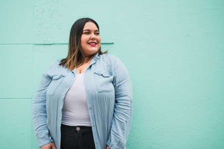 Close-up of young plus size woman smiling while standing against light blue wall outdoors. Urban concept.の写真素材