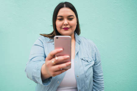 Portrait of young plus size woman doing video call on her mobile phone outdoors against light blue background. Communication concept.の写真素材