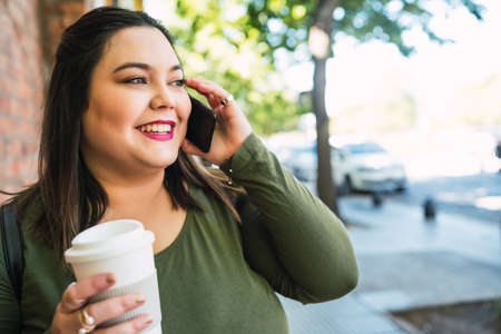 Portrait of young plus size woman talking on the phone while holding a cup of coffee outdoors in the street. Urban concept.の写真素材