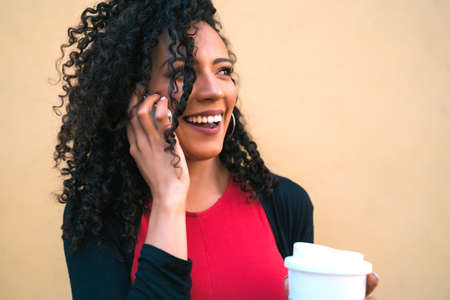Portrait of young afro woman talking on the phone while holding a cup of coffee against yellow background. Communication concept.の写真素材