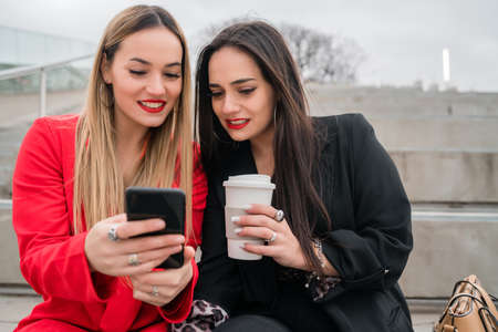 Two friends using their mobile phone while sitting outdoors.