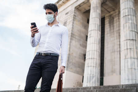 Businessman wearing a face mask and using his mobile phoneの写真素材