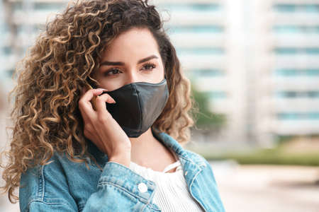 Latin woman using a face mask while talking on the phone outdoors.の写真素材
