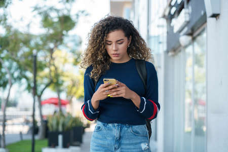 Latin woman using a mobile phone while walking outdoors on the street.の写真素材