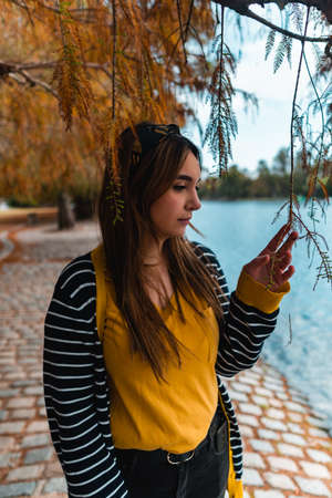 Vertical shot of a young woman touching a hanging leaf at autumnal park with a lakeの写真素材