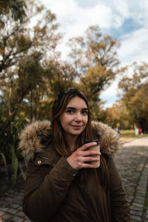 A pretty caucasian woman smiling while holding a coffee and enjoying a walk at park. Verticalの写真素材