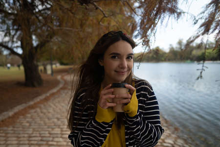 A Pretty woman smiling with a cup of coffee in her hands at autumnal parkの写真素材