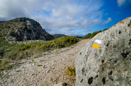 Trail of sort distance in the mountain marked with a sign with two bars white and yellow painted on rock. In the background there is al hill.の写真素材