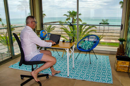Man tele working from home dressed with a elegant shirt for videoconference and shorts and bared feet under the deskの写真素材