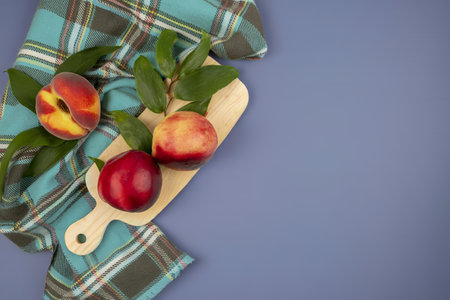 top view of peaches and leaves on cutting board with plaid cloth on blue background with copy space