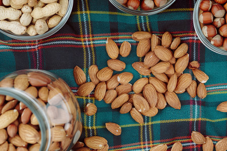 top view of nuts peanuts hazelnuts in bowls and almond scattered from a glass jar on plaid table napkinの写真素材
