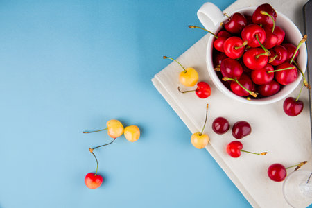 top view of red cherries in cup on cloth and books with yellow cherries