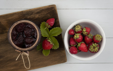 top view of strawberry jam on a glass jar on a wooden kitchen board with fresh strawberries on a white bowlの写真素材