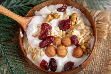 Close-up wooden bowl with yogurt with rolled oats, nuts and dried cranberries and fir twig on burlapの写真素材