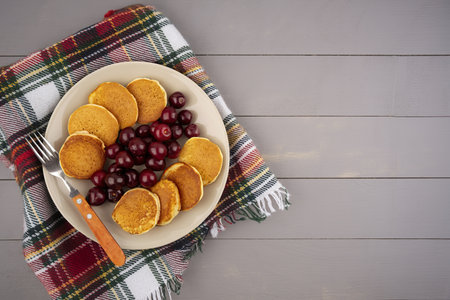 top view of pancakes with cherries and fork in plate on plaid cloth and wooden background with copy spaceの写真素材