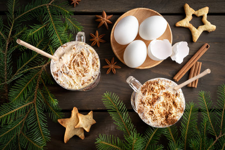 Eggnog with whipped cream in glasses, eggs, whisk, cinnamon, Xmas cookies on old wooden tableの写真素材