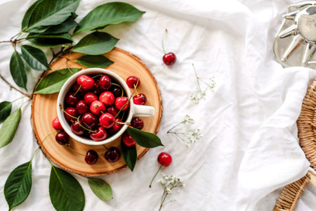 top view of cup full of red cherries on left side and white background decorated with leaves