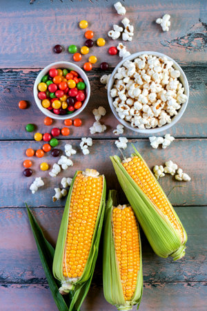 top view of cut corn and uncooked corn with skittles popcorn and corn pop cereal on wooden...